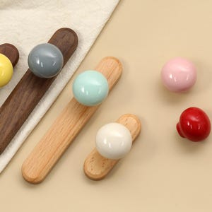 May include: A collection of wooden cabinet knobs with colorful spherical handles. The knobs feature a variety of colors, including yellow, gray, light blue, white, pink, and red. The wooden bases are in shades of brown and light wood.