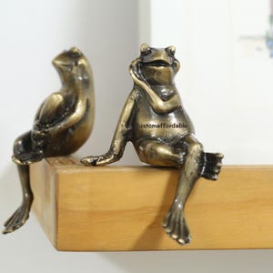 Puede incluir: Dos figuras de rana de bronce sentadas en una estantería de madera. Una rana apoya la barbilla en la mano, mientras que la otra está sentada con las piernas cruzadas. Las ranas tienen un acabado antiguo detallado.