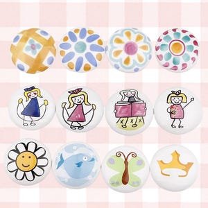 May include: A collection of twelve white ceramic cabinet knobs. The knobs feature hand-painted designs, including floral patterns, children playing, a smiling flower, a fish, a butterfly, and a crown. The knobs are set against a pink and white checkered background.
