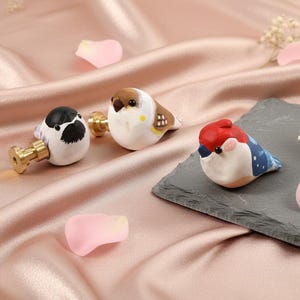 May include: Three ceramic bird figurines in white, brown, black, red, and blue. One bird is mounted on a brass stopper. The birds are on a pink satin surface with scattered rose petals and sprigs of white flowers.