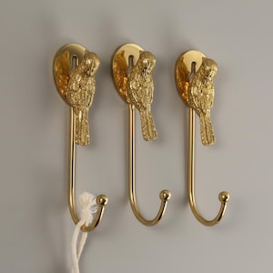 May include: Three gold-coloured metal wall hooks. Each hook features a decorative parrot design at the top and a curved hook at the bottom. The hooks are mounted on a light grey wall.