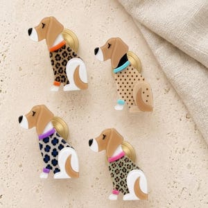 May include: Four dog-shaped cabinet knobs featuring tan heads and white accents. Each knob displays a different patterned sweater: one with brown leopard print, one with brown polka dots, one with blue floral shapes, and one with green leopard print.