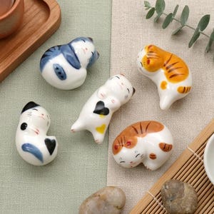 以下が含まれることがあります： 様々なポーズの猫の形をしたセラミック箸置きのコレクション。猫は白で、青、オレンジ、茶色の模様があります。箸置きは、小さな白い皿と木製のトレイとともに、薄緑色とベージュ色の表面に配置されています。