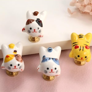 Puede incluir: Un conjunto de cinco pomos de cerámica con forma de gato. Los pomos presentan varios colores y diseños, incluyendo blanco, amarillo, marrón y azul, con bases doradas. Los pomos de gato están diseñados con una estética caprichosa y de dibujos animados.