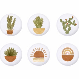 May include: Six round white buttons with different illustrations. The buttons feature a cactus in a pot, a sun, a rainbow, and three different types of cacti.