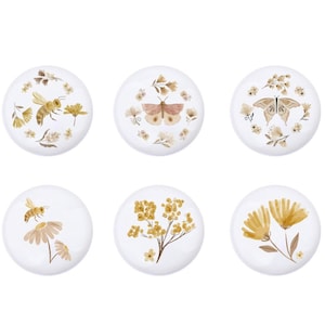 May include: Six round white coasters featuring various floral and insect designs. The designs include a bee, a butterfly, a moth, and different floral arrangements in shades of brown and yellow.