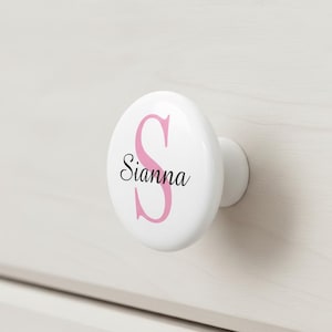 May include: White ceramic drawer knob with a pink letter "S" and the name "Sianna" in black script. The knob is mounted on a white wooden surface. A decorative accent for furniture.