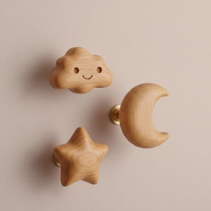 Puede incluir: Tres tiradores de armario de madera con forma de nube, estrella y luna creciente. La nube tiene una cara sonriente tallada. Los tiradores son de color marrón claro y están montados sobre herrajes de latón, sobre un fondo neutro.
