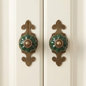 May include: Two cabinet knobs with a dark green, flower-shaped ceramic handle and a brass center. The knobs are mounted on ornate, bronze-colored backplates. The cabinet doors are painted white.