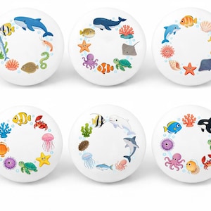 May include: Six white ceramic cabinet knobs with colorful sea creature designs. Each knob features a different arrangement of marine life, including whales, dolphins, fish, and coral, creating a playful ocean theme.