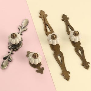 May include: A collection of antique-style cabinet hardware. The hardware features white ceramic flower-shaped knobs with bronze-colored metal bases and pulls. The text "customaffordable" is printed on one of the pulls.