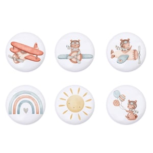 Könnte beinhalten: Sechs runde, weiße, illustrierte Knöpfe mit verschiedenen Designs. Die Designs beinhalten ein rotes Flugzeug, einen Bären in einem Flugzeug, einen Regenbogen, eine Sonne, einen Bären mit Ballons und einen Bären in einem Flugzeug.