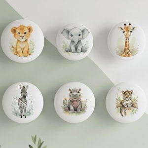 May include: Six white ceramic cabinet knobs featuring watercolour illustrations of safari animals: a lion cub, elephant, giraffe, zebra, hippo, and leopard. Each knob is round with a detailed animal design.