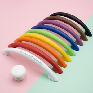 Peut inclure: Une collection de poignées de meubles colorées et un bouton blanc, disposées sur un fond rose et vert. Les poignées sont de différentes couleurs, dont le rouge, l'orange, le jaune, le vert, le bleu, le violet, le marron et le noir. La poignée blanche porte le texte "Customaffordable".