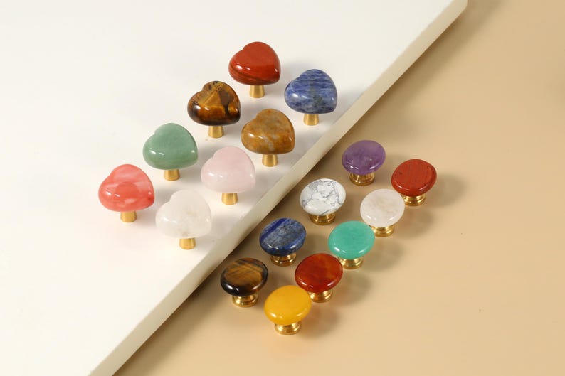 May include: A selection of heart-shaped and round cabinet knobs in a variety of colours and materials, including red, green, blue, and white. Each knob has a gold-coloured base. The knobs are made of stone and are arranged on a white surface.
