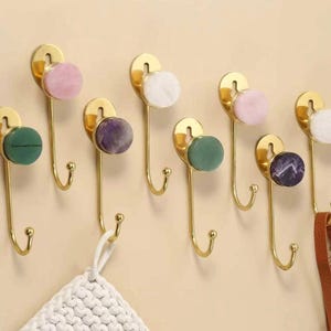 May include: A collection of decorative wall hooks with brass-colored metal and colorful, round stone accents. The hooks are mounted on a light-colored wall, with a few items hanging from them, including a white crocheted item and a brown leather strap.