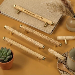 Pode incluir: Uma coleção de puxadores de armário em rattan tecido com ferragens em latão. Os puxadores são cilíndricos e de cor bege natural, dispostos sobre uma superfície neutra. Um dos puxadores tem o texto "Customaffordable" impresso. Uma planta suculenta num vaso de terracota está em primeiro plano.
