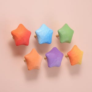 May include: Six colorful star-shaped cabinet knobs in shades of orange, blue, green, purple, and yellow. Each knob has a gold-colored base. The knobs are arranged on a light pink background.