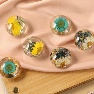 Puede incluir: Seis esferas de resina transparente, cada una encapsulando una flor vibrante y hojuelas doradas. Las flores son amarillas, azules y blancas. Las esferas están dispuestas sobre una superficie de madera y tela color melocotón.