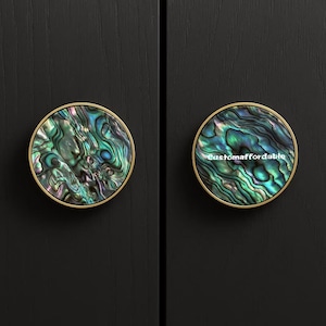 Könnte beinhalten: Zwei runde Schrankknöpfe mit schillernden Abalone-Muschel-Mittelstücken und goldfarbenen Rändern. Die Knöpfe sind vor einem dunklen Hintergrund angeordnet. Ein Knopf trägt den Text "customaffordable".