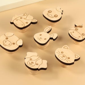 May include: Six wooden knobs featuring various animal faces. The knobs are light brown with a dark brown outline. The animals depicted are a pig, a cow, a chicken, a rabbit, a cat, and a dog.