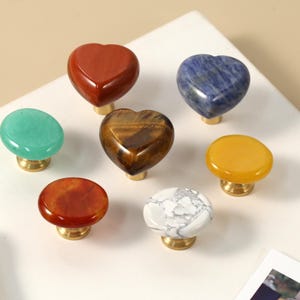 May include: A selection of gemstone cabinet knobs in different shapes and colours. The knobs include heart-shaped red jasper, blue sodalite, and a tiger's eye heart. Other knobs are green, orange, yellow, and white with grey veining. Each knob has a brass base.