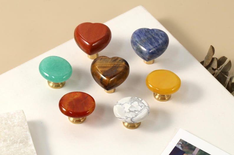 May include: A selection of gemstone cabinet knobs in different shapes and colours. The knobs include heart-shaped red jasper, blue sodalite, and a tiger's eye heart. Other knobs are green, orange, yellow, and white with grey veining. Each knob has a brass base.