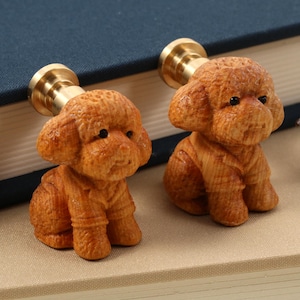 May include: Two wooden dog-shaped cabinet knobs with brass hardware. The knobs are carved in detail, depicting a seated poodle. The wood has a warm, reddish-brown tone. The knobs are placed on a neutral surface next to a book.