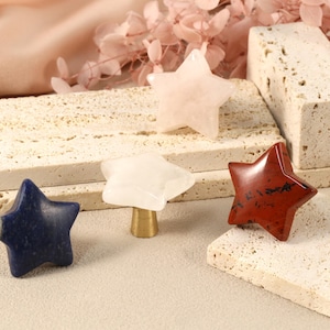 May include: Five star-shaped drawer pulls made of different types of stone. The pulls are in shades of pink, white, blue, and red. The pulls are on a light beige surface.