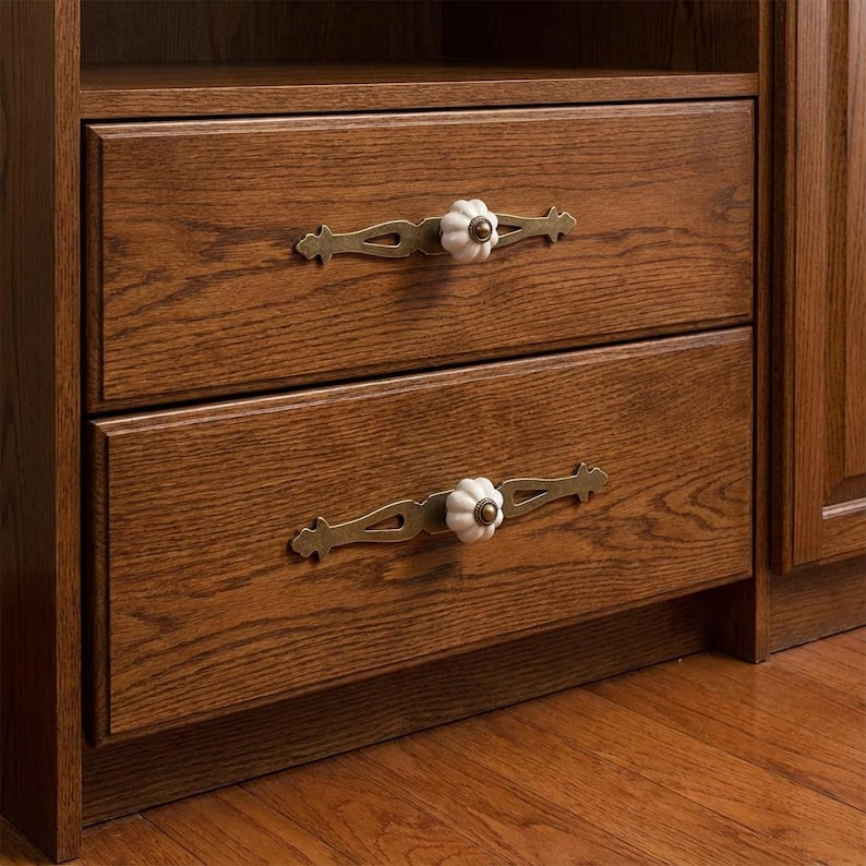 May include: Close-up of a wooden cabinet with two drawers. Each drawer has a decorative brass handle with a white ceramic flower-shaped knob. The wood has a rich, warm brown tone.