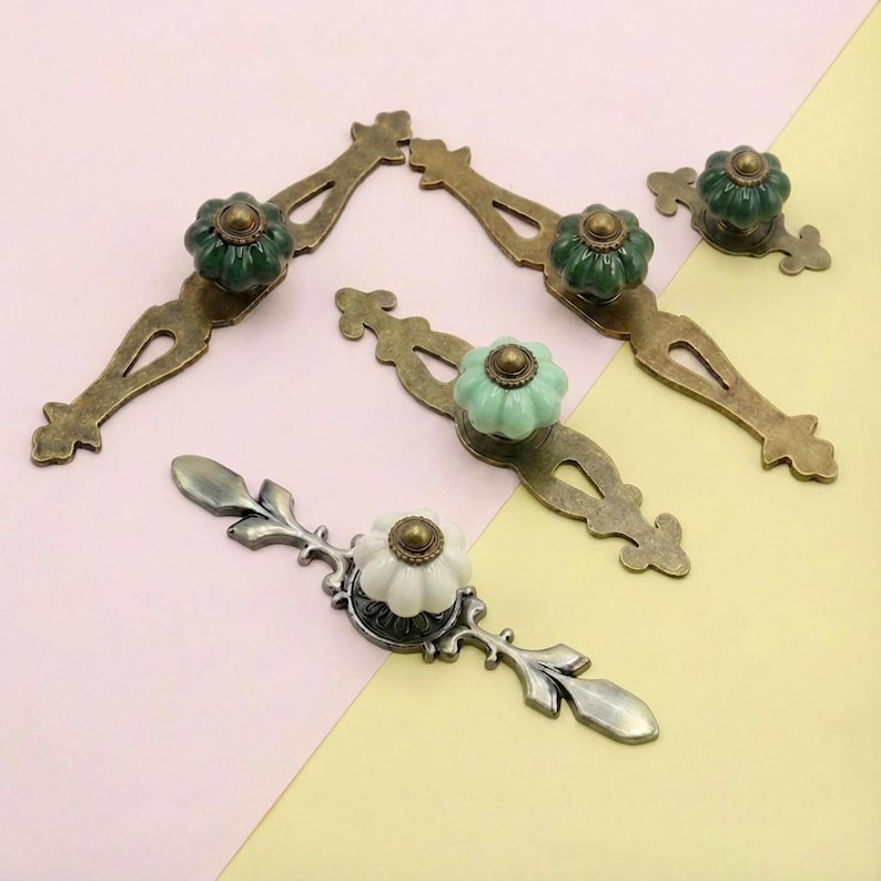 May include: A collection of decorative cabinet pulls with ornate, antique-style designs. The pulls feature brass-toned metal bases and ceramic knobs in green, white, and light green. The knobs have a floral or pumpkin-like shape.