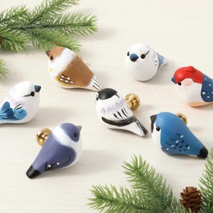 Puede incluir: Una colección de siete pomos decorativos con forma de pájaro en varios colores, incluyendo azul, marrón y blanco. Cada pomo presenta una base dorada y está diseñado para parecerse a una especie de ave diferente. Ramas de pino verdes y una piña en el fondo.
