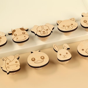 May include: A collection of ten wooden cabinet knobs, each designed with a different animal face. The knobs feature a light wood color with dark brown outlines, depicting a pig, cow, dog, cat, duck, horse, and rabbit.