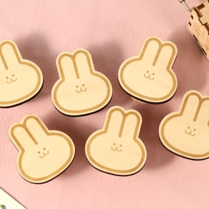 May include: Six wooden bunny-shaped ornaments with a light brown finish. Each ornament has a simple, smiling face.