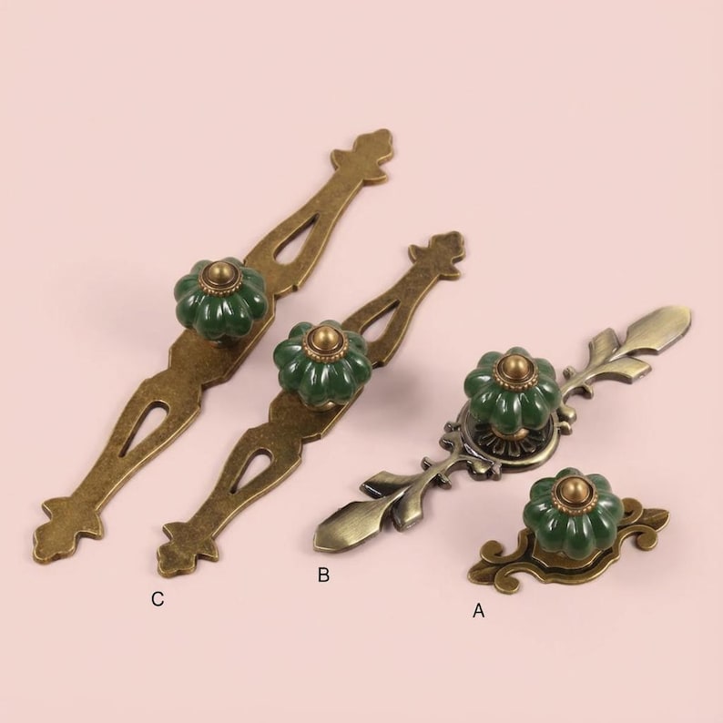 May include: A collection of antique-style cabinet hardware. The hardware features green ceramic knobs with a flower design, set on bronze-colored metal backplates. The backplates have ornate, vintage-inspired shapes, including pulls and knobs.