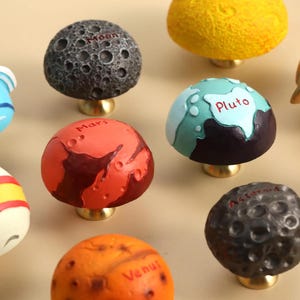 May include: A collection of decorative cabinet knobs, each designed to resemble a planet or celestial body. The knobs feature various colors and textures, with names like "Uranus," "Mars," and "Pluto" written on them. They have brass-colored bases.