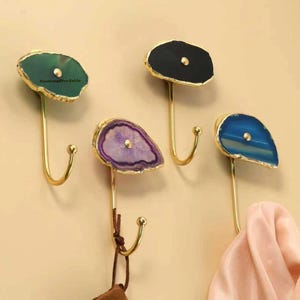 Könnte beinhalten: Vier dekorative Wandhaken mit Oberteilen aus Achatstein in Grün, Schwarz, Lila und Blau. Jeder Haken hat einen goldfarbenen Metallstiel und einen gebogenen Haken zum Aufhängen von Gegenständen. Ein rosa Schal hängt an einem Haken.