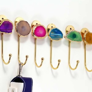 Peut inclure: Six crochets muraux décoratifs avec une quincaillerie en métal doré. Chaque crochet présente une pierre d'agate colorée unique dans les tons de violet, marron, rose, bleu, vert et jaune. Un étui bleu et blanc est suspendu à un crochet.
