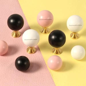 以下が含まれることがあります： 黒、白、ピンクの装飾的なキャビネットノブのコレクション。ノブは球形デザインで、金色のベースに取り付けられています。一部のノブには「customaffordable」の文字が印刷されています。背景はピンクと黄色に分かれています。