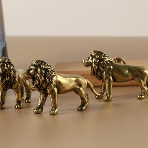 Op de afbeelding: Drie gouden leeuwenbeeldjes, elk in een andere pose. De leeuwen staan op een lichtbruin oppervlak.