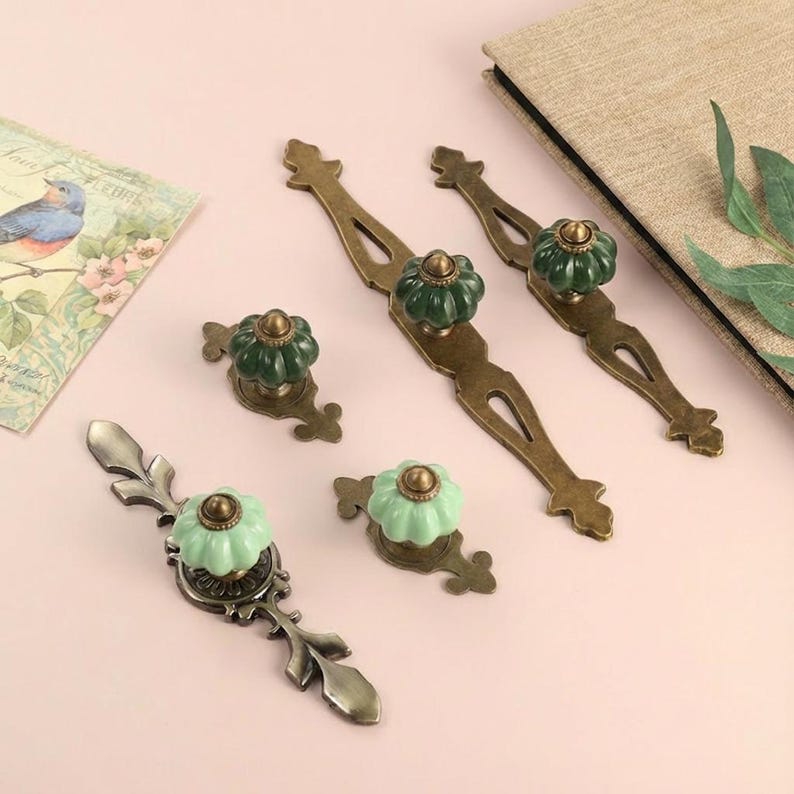 May include: A collection of antique-style cabinet hardware, including drawer pulls and knobs. The pulls have a bronze finish with decorative cutouts, and the knobs feature green and light green ceramic flower-shaped designs. The hardware is arranged on a pink surface.