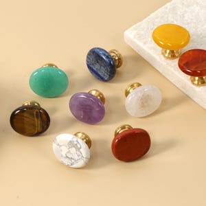 May include: A selection of gemstone cabinet knobs with brass fittings. The knobs are in various shapes and colours, including green, blue, purple, white, red, and yellow. The knobs are displayed on a neutral background.