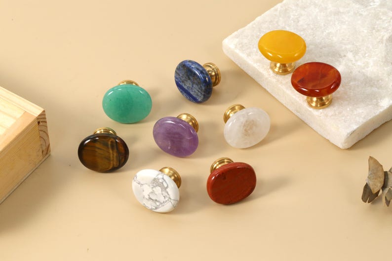 May include: A selection of gemstone cabinet knobs with brass fittings. The knobs are in various shapes and colours, including green, blue, purple, white, red, and yellow. The knobs are displayed on a neutral background.