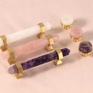May include: A selection of crystal cabinet pulls and knobs. The pulls are white, pink, and purple, with gold-coloured metal hardware. The knobs are white, pink, and purple with gold-coloured metal bases. They are arranged on a light pink surface.