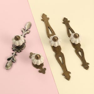 May include: A collection of antique-style cabinet hardware. The set includes two long pulls, two smaller pulls, and two knobs. Each piece features a white ceramic flower-shaped knob with a brass center, set against a bronze-colored metal base.