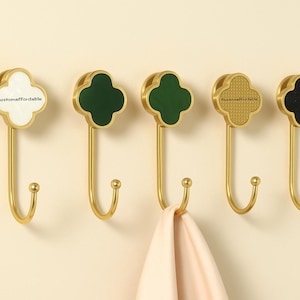 Può includere: Cinque ganci da parete color oro con un design a trifoglio. I ganci hanno centri a trifoglio di diversi colori: bianco, verde scuro, verde chiaro, oro e nero. Un panno rosa chiaro è appeso a un gancio. Il testo "customaffordable" è sul gancio bianco.