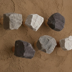 Może przedstawiać: Sześć dekoracyjnych gałek w kształcie skał w odcieniach szarości i czerni, zamontowanych na drewnianych podstawach. Gałki są przymocowane do teksturowanej brązowej powierzchni, oferując naturalną, ziemistą estetykę. Idealne do szafek lub szuflad.