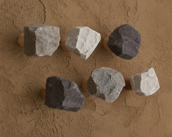 May include: Six decorative rock knobs in shades of gray and black are mounted on wooden bases. The knobs are affixed to a textured brown surface, offering a natural, earthy aesthetic. These knobs are ideal for cabinets or drawers.