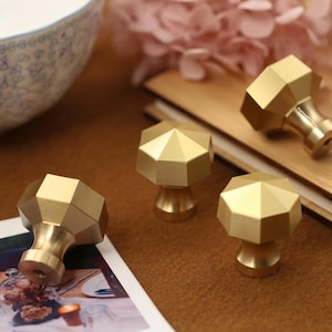 以下が含まれることがあります： 茶色の表面に配置された、ファセットデザインの4つの金色の幾何学的なキャビネットノブ。ノブは円筒形のベースと多面的な上部を持っています。部分的に見えるボウルと写真が背景にあります。