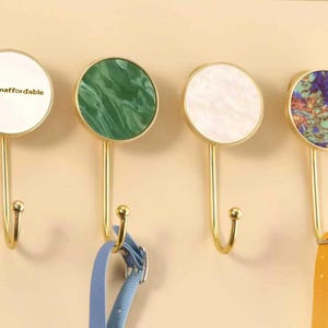 Puede incluir: Cuatro ganchos decorativos de pared con tallos dorados y tapas circulares. Las tapas presentan diferentes diseños: blanco con texto, mármol verde, mármol blanco y un patrón colorido. Una correa azul y otra amarilla cuelgan de dos de los ganchos.